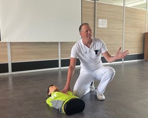 Ein Mann in weißer Kleidung demonstriert Erste Hilfe an einer Übungspuppe, die auf dem Boden liegt.