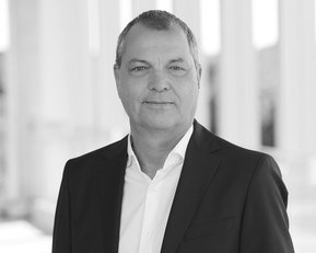 Mann in Anzug steht vor einer hellen, modernen Architektur. Er hat kurze Haare und schaut direkt in die Kamera.