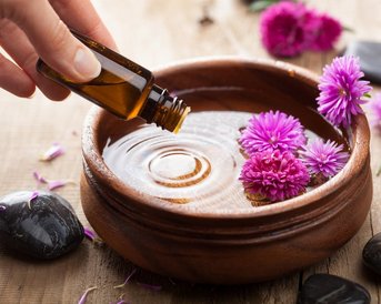 Eine Hand hält eine Flasche ätherisches Öl über eine Schüssel mit Wasser, in der lila Blumen schwimmen. Rund um die Schüssel liegen schwarze Steine und einige Blütenblätter.