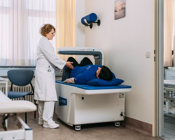 orthopaedie Eine medizinische Fachkraft hilft einem Patienten, der auf einer Behandlungsliege liegt, während eine Therapieeinheit vorbereitet wird.