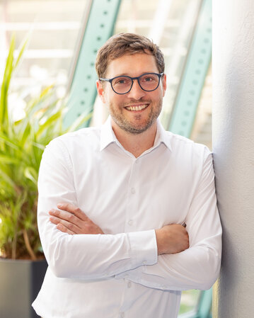 Ein Mann mit Brille in einem weißen Hemd lehnt an einer Säule, umgeben von Pflanzen in einem modernen Raum.