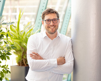 Ein Mann mit Brille in einem weißen Hemd lehnt an einer Säule, umgeben von Pflanzen in einem modernen Raum.